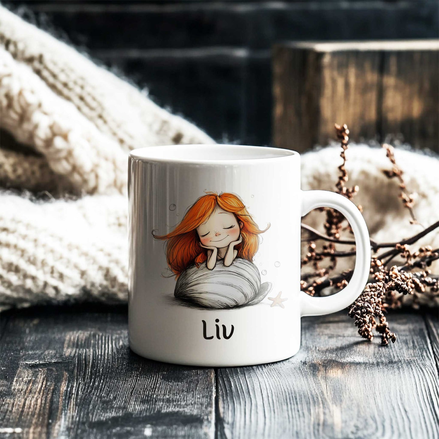 Witte keramische mok met een zeemeermin met lang oranje haar op een grote witte schelp. De mok staat op een donkere houten tafel naast een zachte gebreide deken. Onder de illustratie staat de naam Liv.