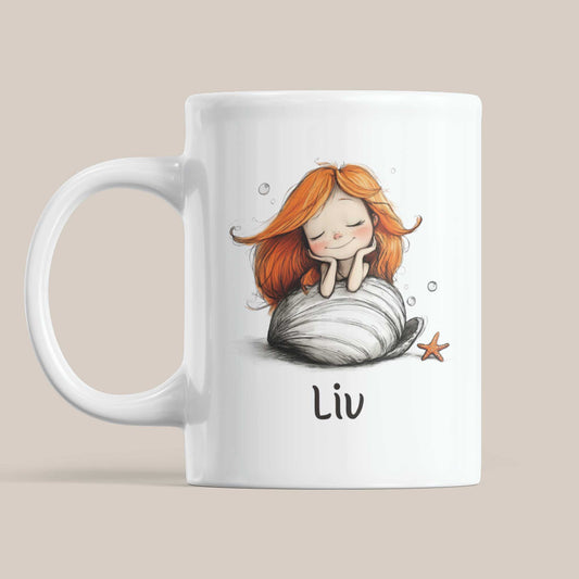 Witte keramische mok met een illustratie van een dromende zeemeermin met lang oranje haar op een grote witte schelp. Onder de illustratie staat de naam Liv. op beige achtergrond