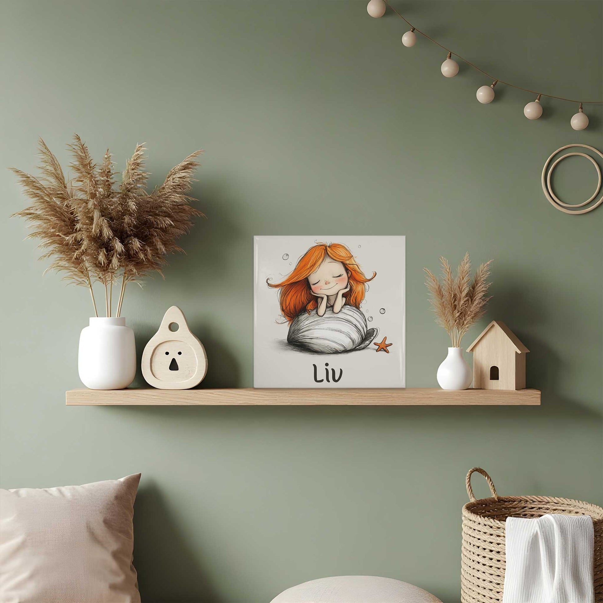 Keramisch tegeltje met een zeemeermin met lang oranje haar die dromend op een grote witte schelp rust. Onder de illustratie staat de naam Liv. Het tegeltje staat op een houten plank tegen een groene muur.