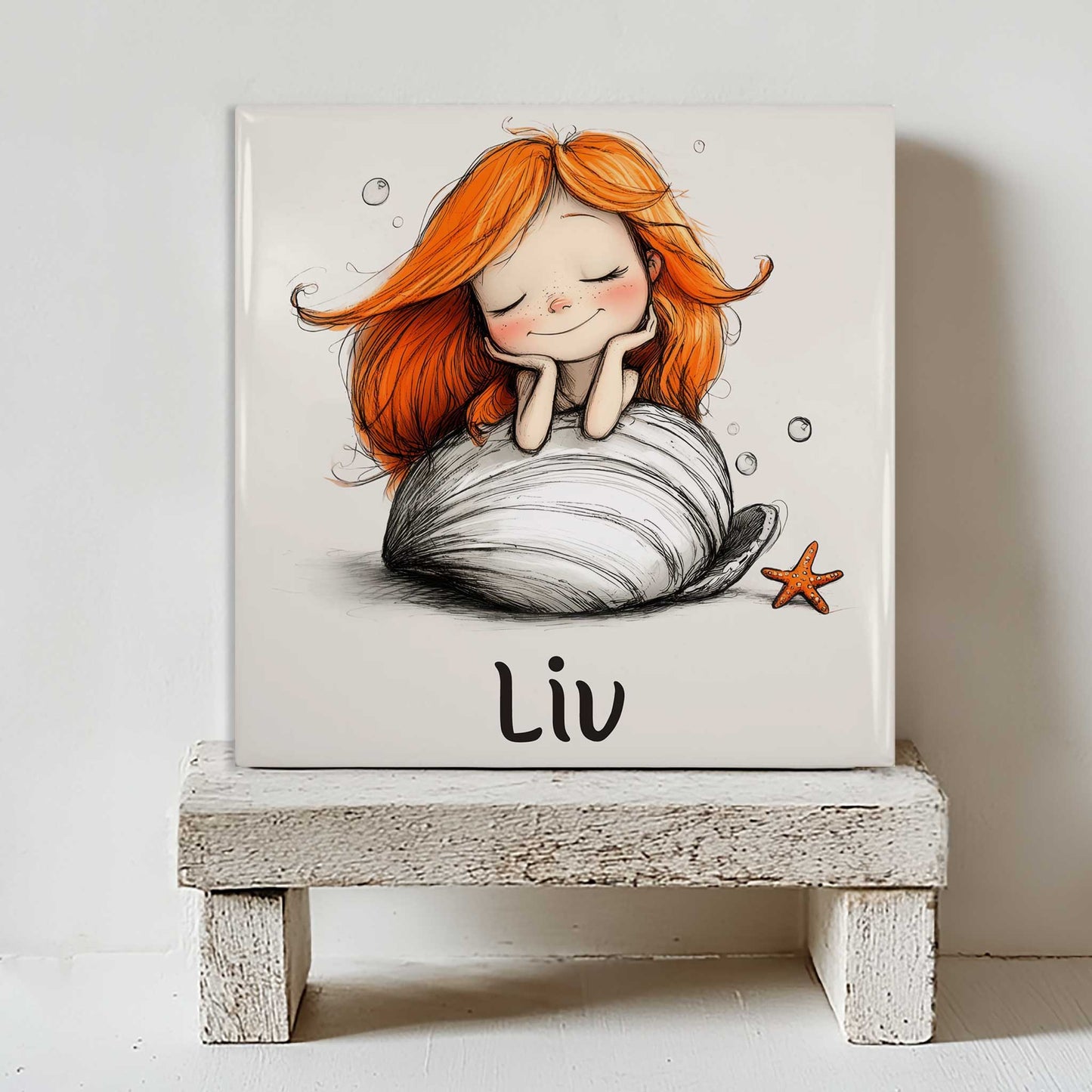 Keramisch tegeltje met een zeemeermin met lang oranje haar op een grote witte schelp. Het tegeltje staat op een klein wit houten krukje. Onder de illustratie staat de naam Liv.