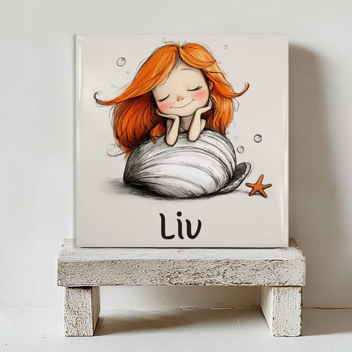 Keramisch tegeltje met de naam Liv en een dromende zeemeermin met oranje haar op een grote schelp. Het tegeltje staat op een wit houten krukje in een lichte kinderkamer.