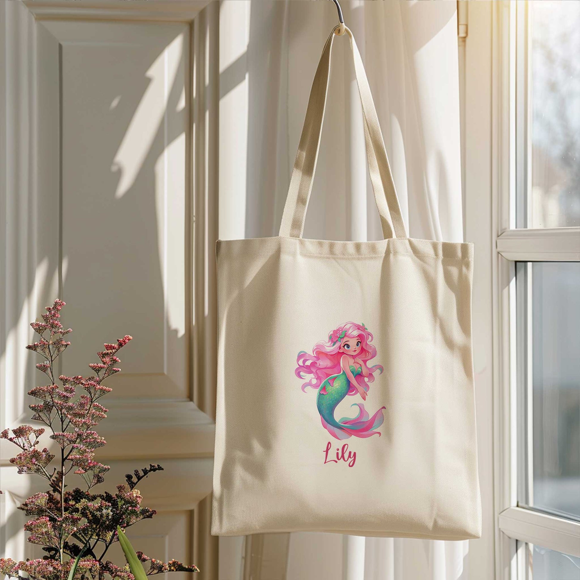 Gepersonaliseerde totebag hangend bij een raam. Op de tas staat een zeemeermin met roze haar, bloemen in haar haar en een kleurrijke turquoise zeemeerminstaart. Onder de illustratie staat de naam Lily.