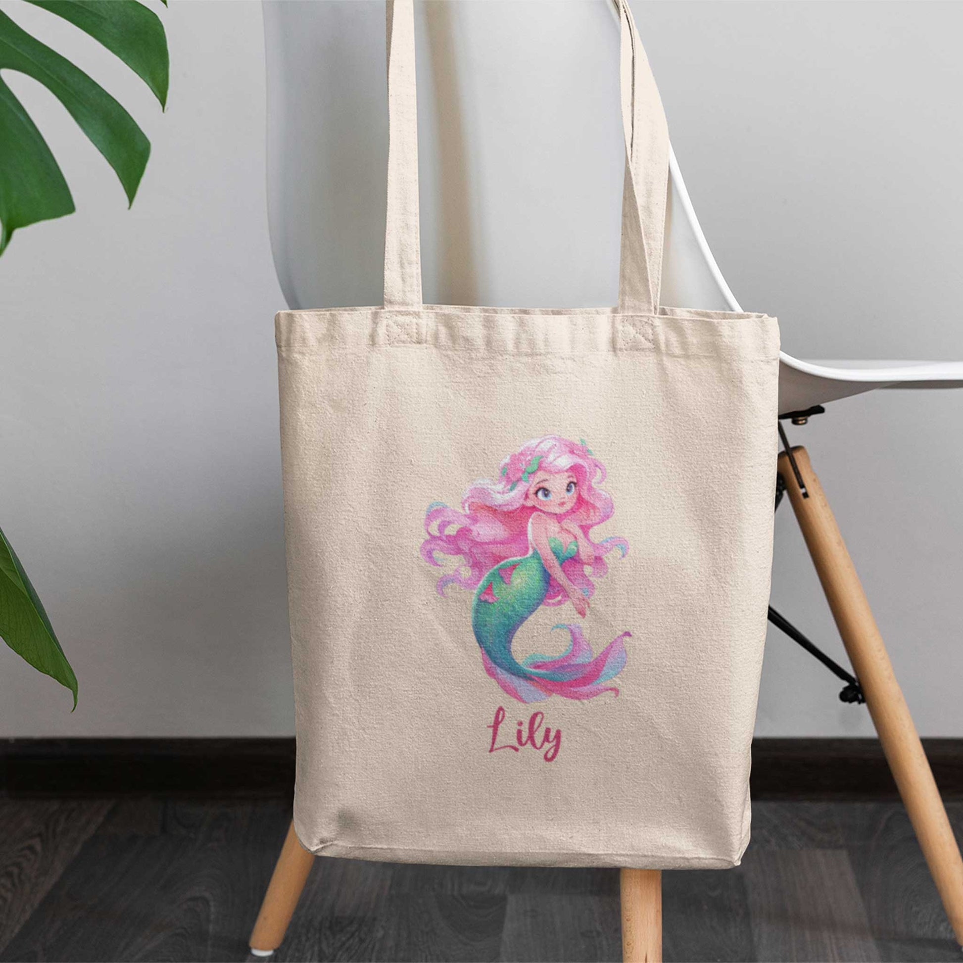 Katoenen totebag met de naam Lily. Op de tas staat een vrolijke zeemeermin met lang golvend roze haar, bloemen in haar haar, grote blauwe ogen en een turquoise zeemeerminstaart met roze accenten. Tas hangt aan stoel.