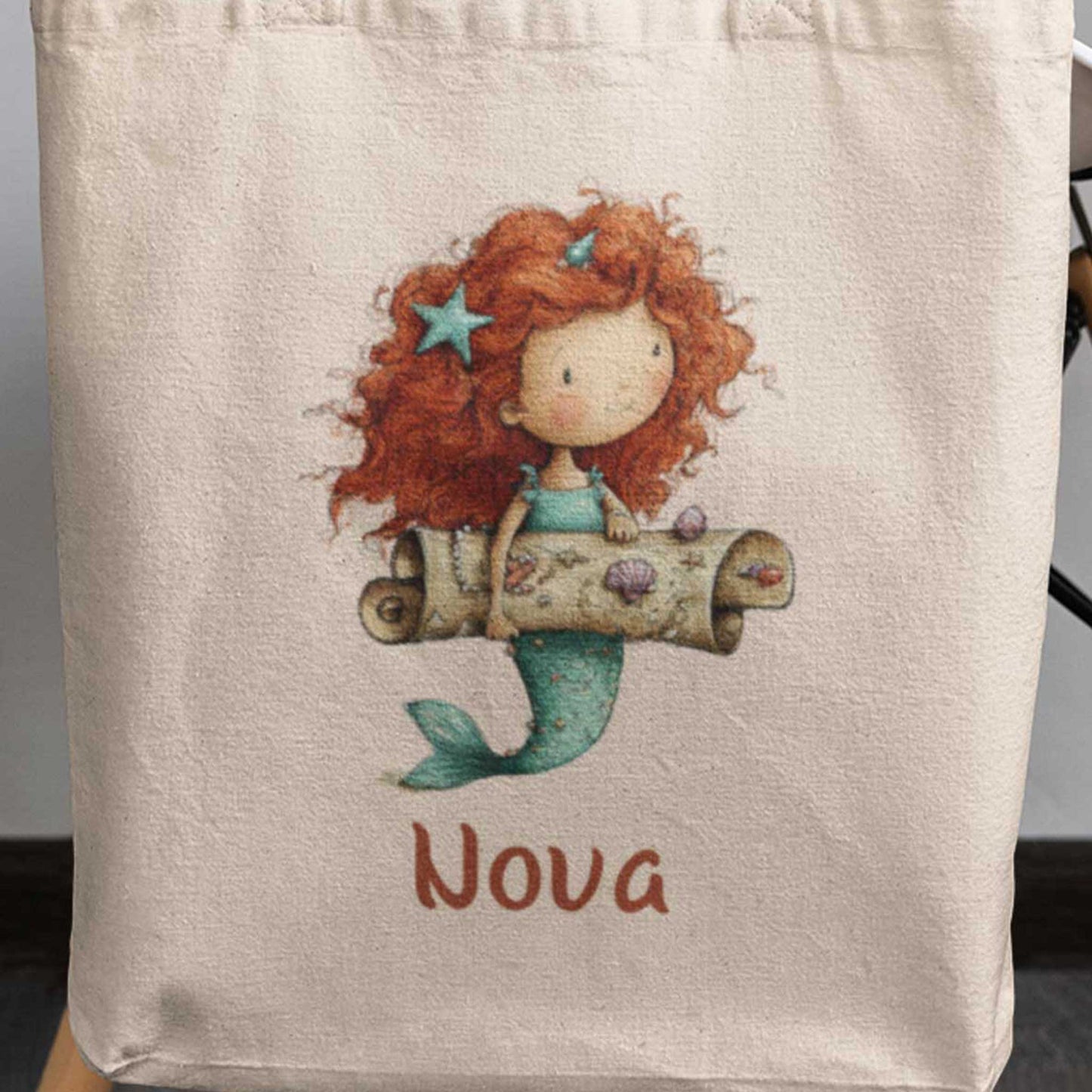 Close-up van katoenen totebag met naam Nova en illustratie van een zeemeermin met schatkaart en schelpen.