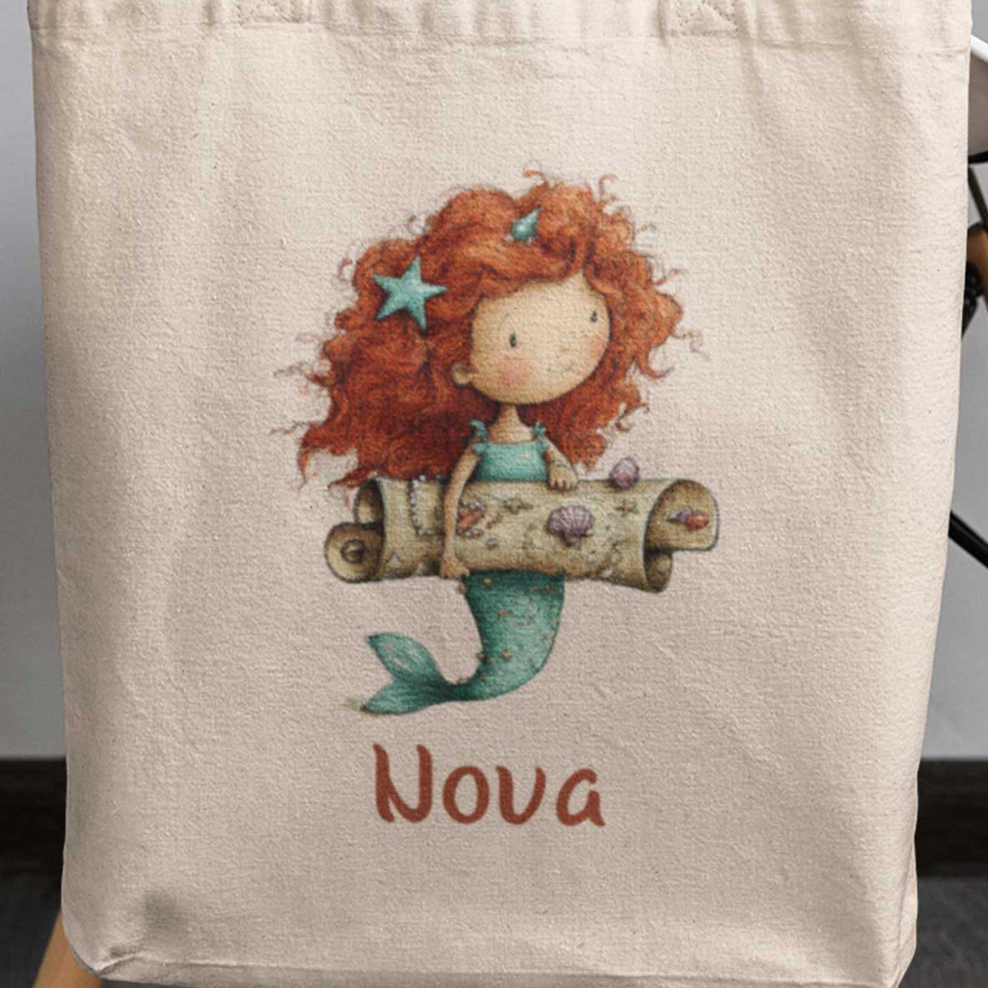 Close-up van katoenen totebag met naam Nova en illustratie van een zeemeermin met schatkaart en schelpen.