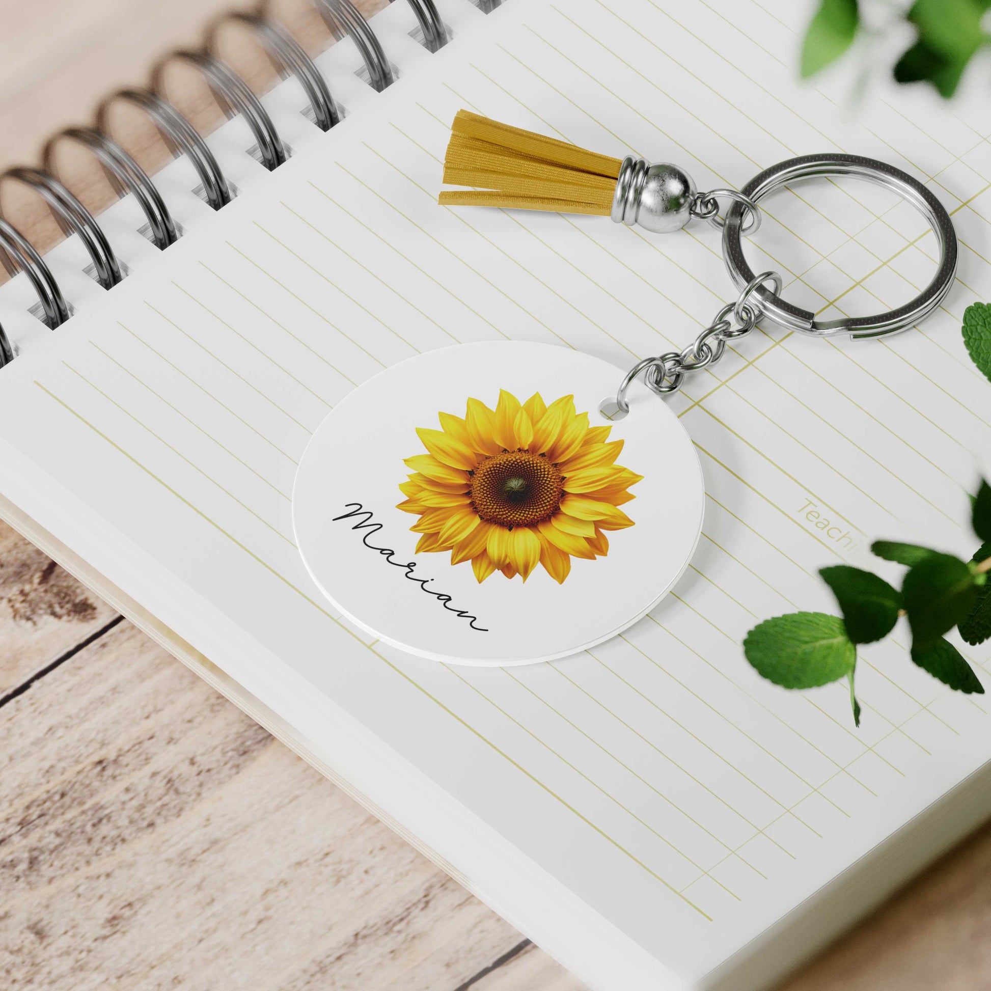 Ronde sleutelhanger met een zonnebloem en de naam Marian ligt op een open notitieboek naast een sleutelbos met een gele tassel.