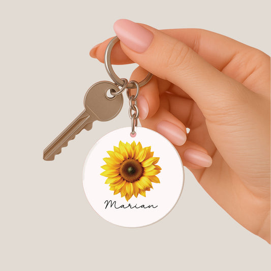 Een vrouw houdt een ronde sleutelhanger met een gele zonnebloem en de naam Marian tussen haar vingers vast. Aan de metalen ring hangt één zilverkleurige sleutel.