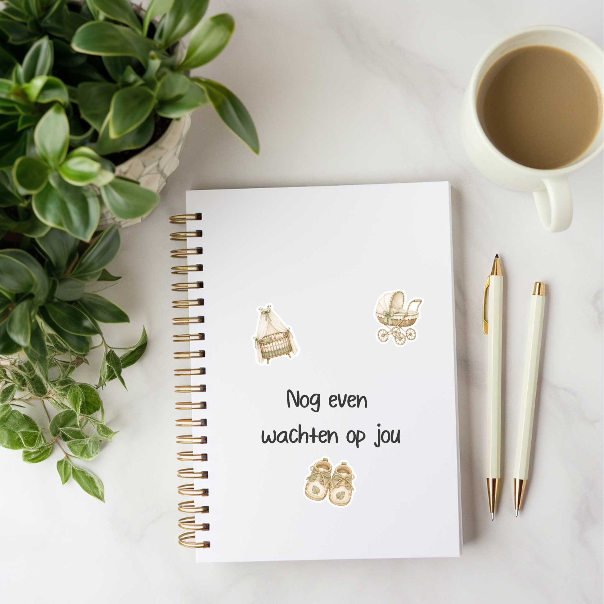 Wit notitieboek met de tekst 'Nog even wachten op jou', versierd met safari baby stickers van een wiegje, kinderwagen en babyschoentjes, liggend op een bureau naast groene planten, een pen en een kop koffie.