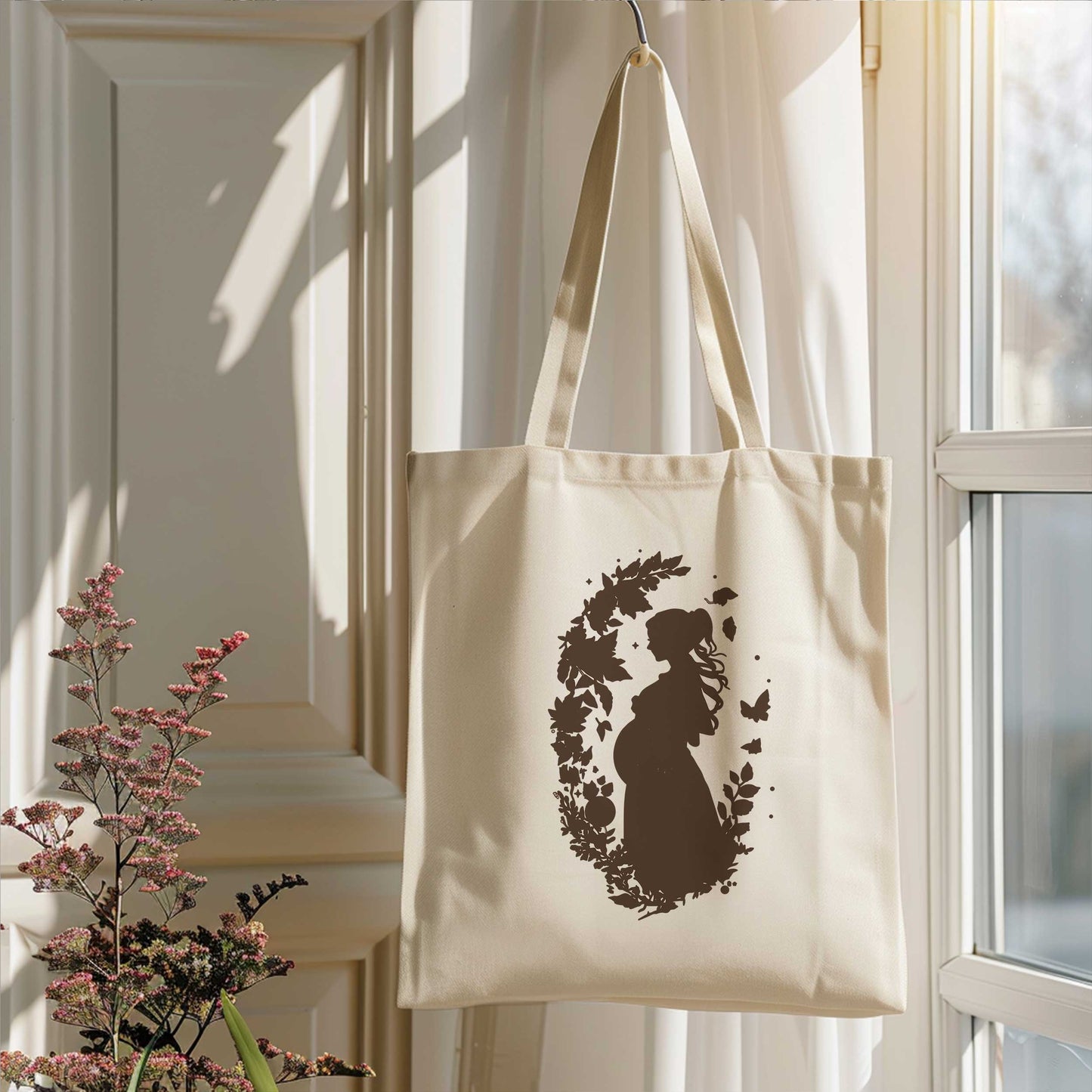 Crèmekleurige totebag met zwangerschapsillustratie hangend naast een raam in een licht interieur met bloemen.
