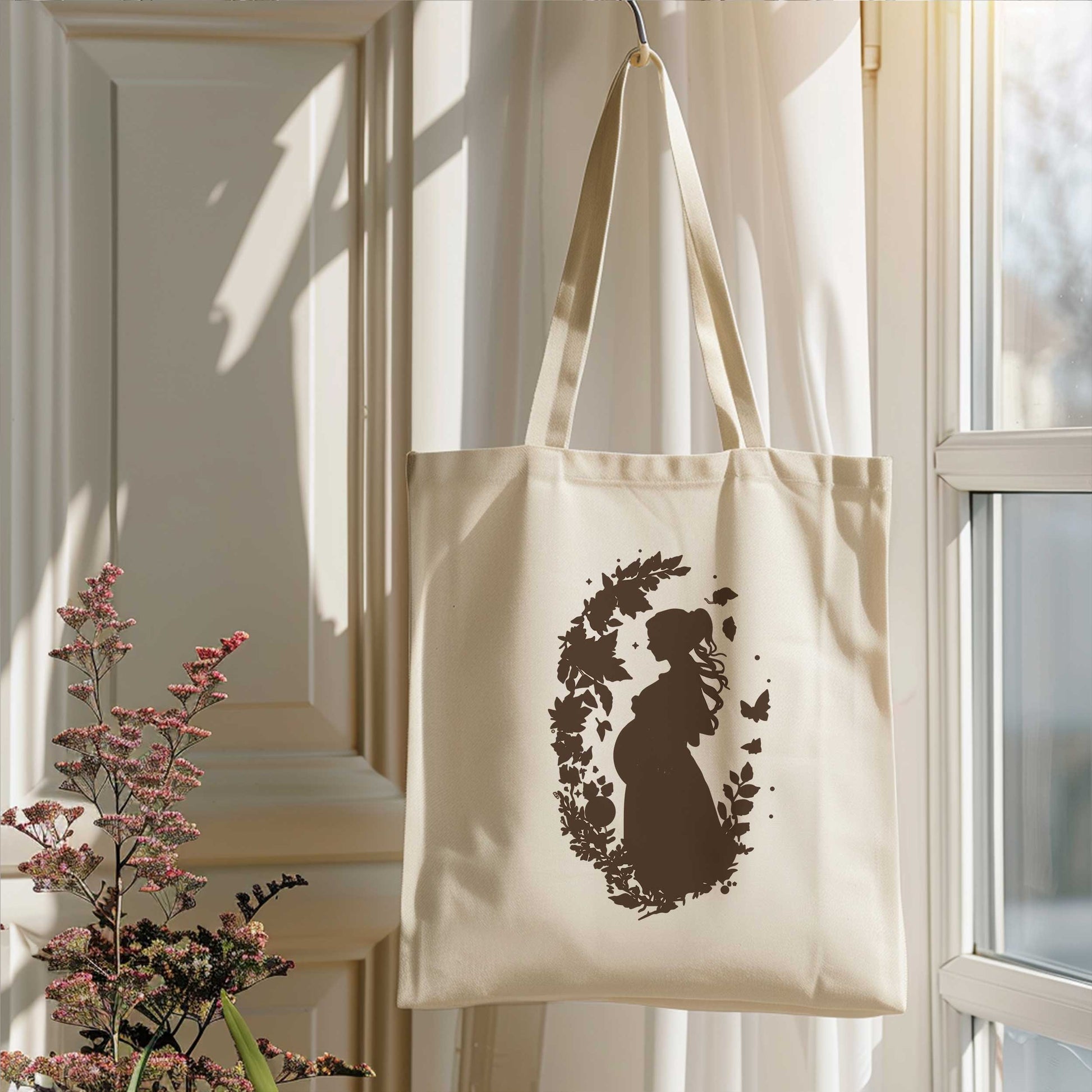Crèmekleurige totebag met zwangerschapsillustratie hangend naast een raam in een licht interieur met bloemen.