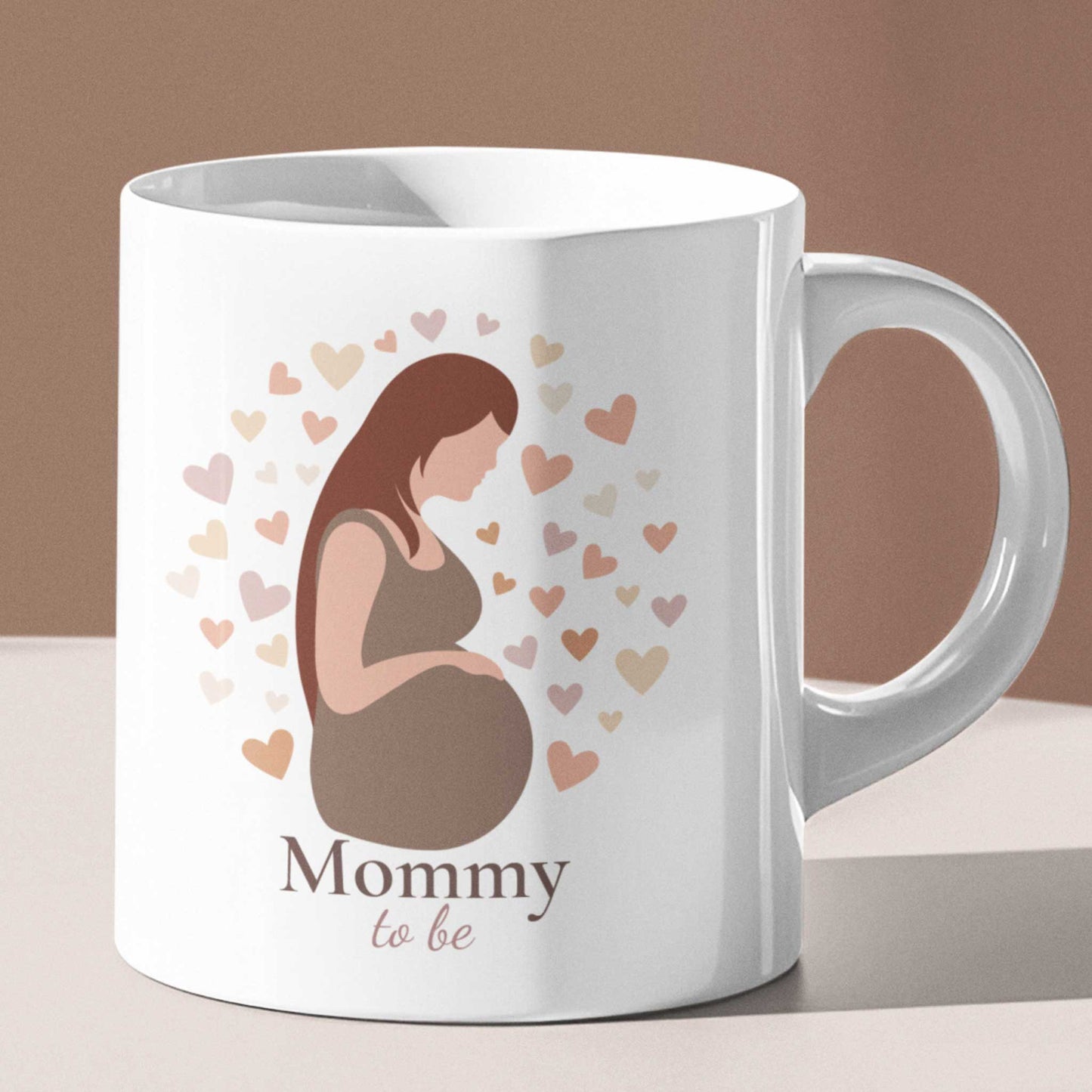 Witte zwangerschapsmok met Mommy To Be illustratie wordt vastgehouden door een vrouw met gouden sieraden tegen een lichte achtergrond.